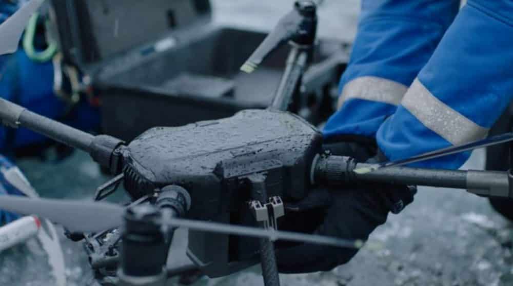 A worker in blue overalls holds a rain-soaked industrial quadcopter outdoors.