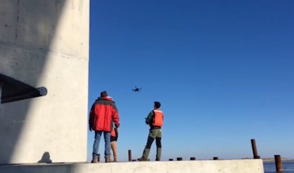 Two inspectors on a concrete bridge pier watch a small drone fly over the water.