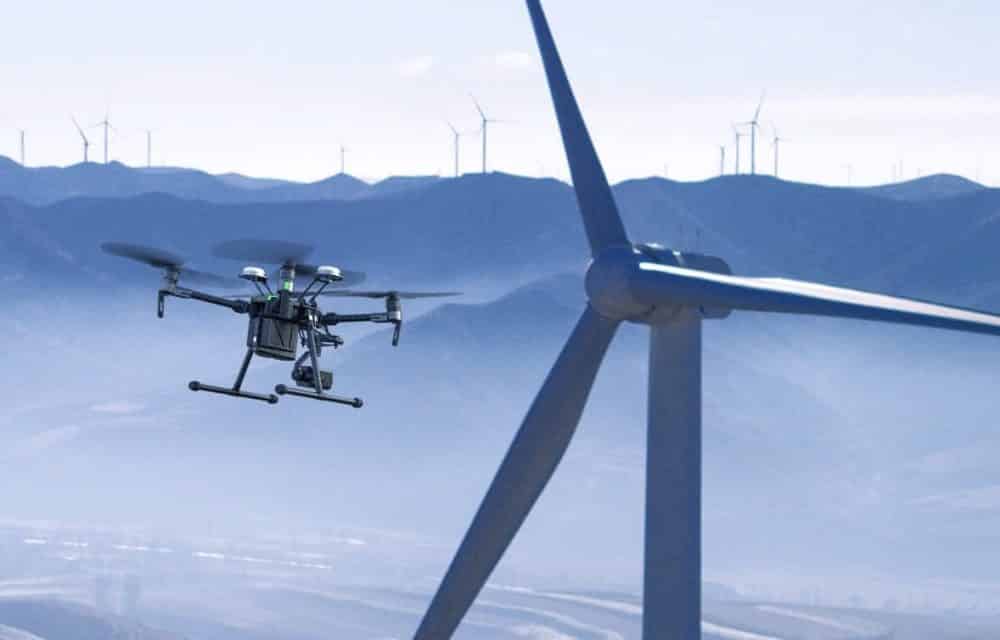 Enterprise drone inspects a wind turbine blade with a mountain wind farm behind.