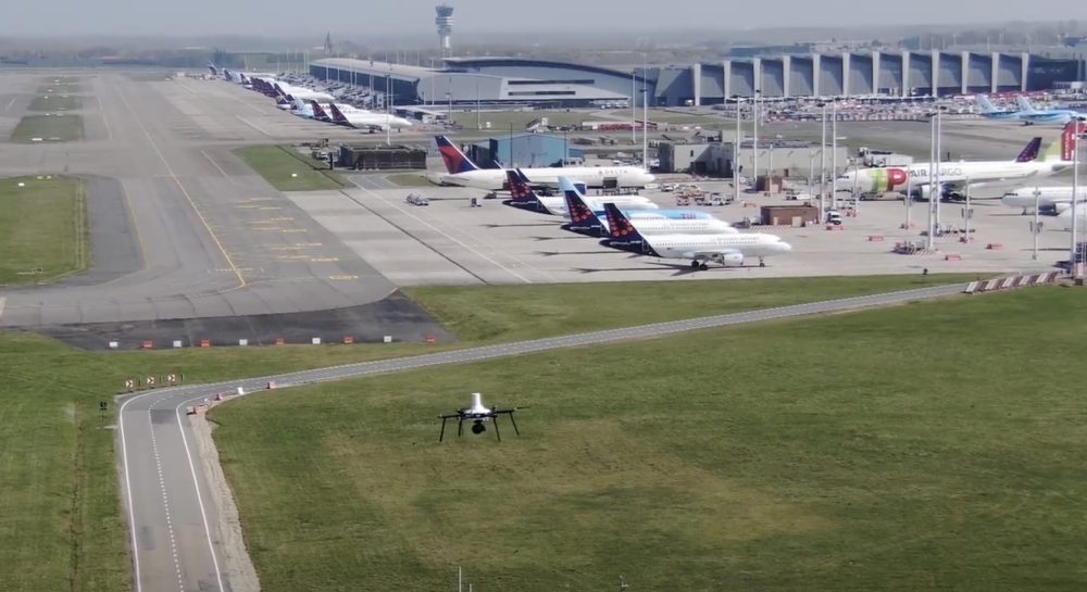 Small drone hovering over grass beside the taxiway at Brussels Airport.