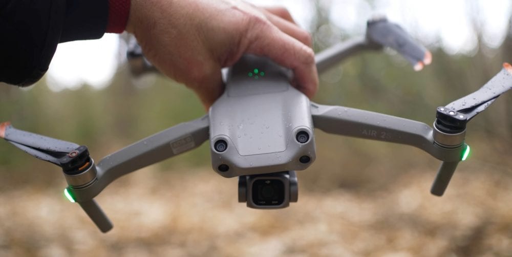 A hand holds a hovering DJI Air 2S by its body above the forest floor.