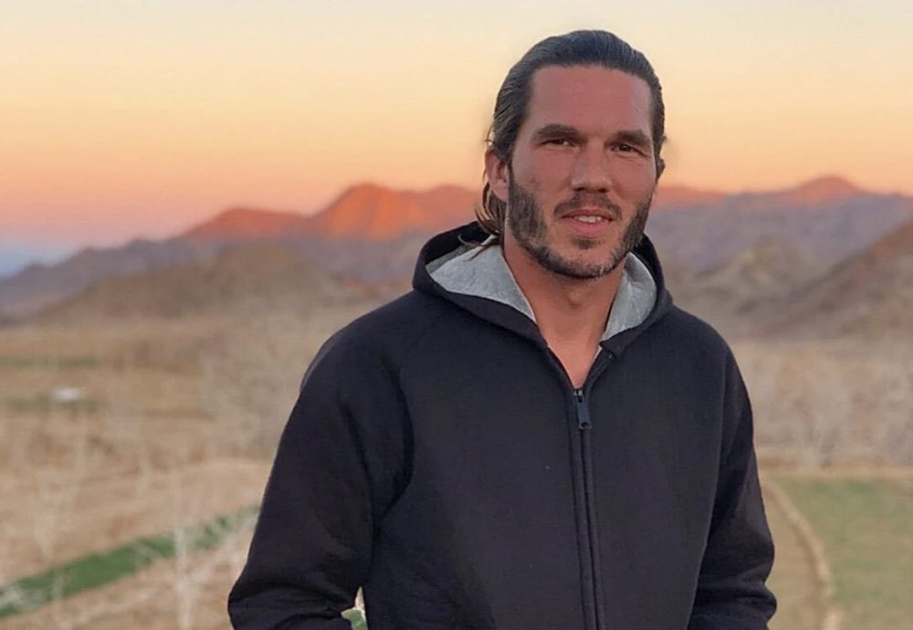 Portrait of a young man standing outdoors against a mountain ridge at dusk.
