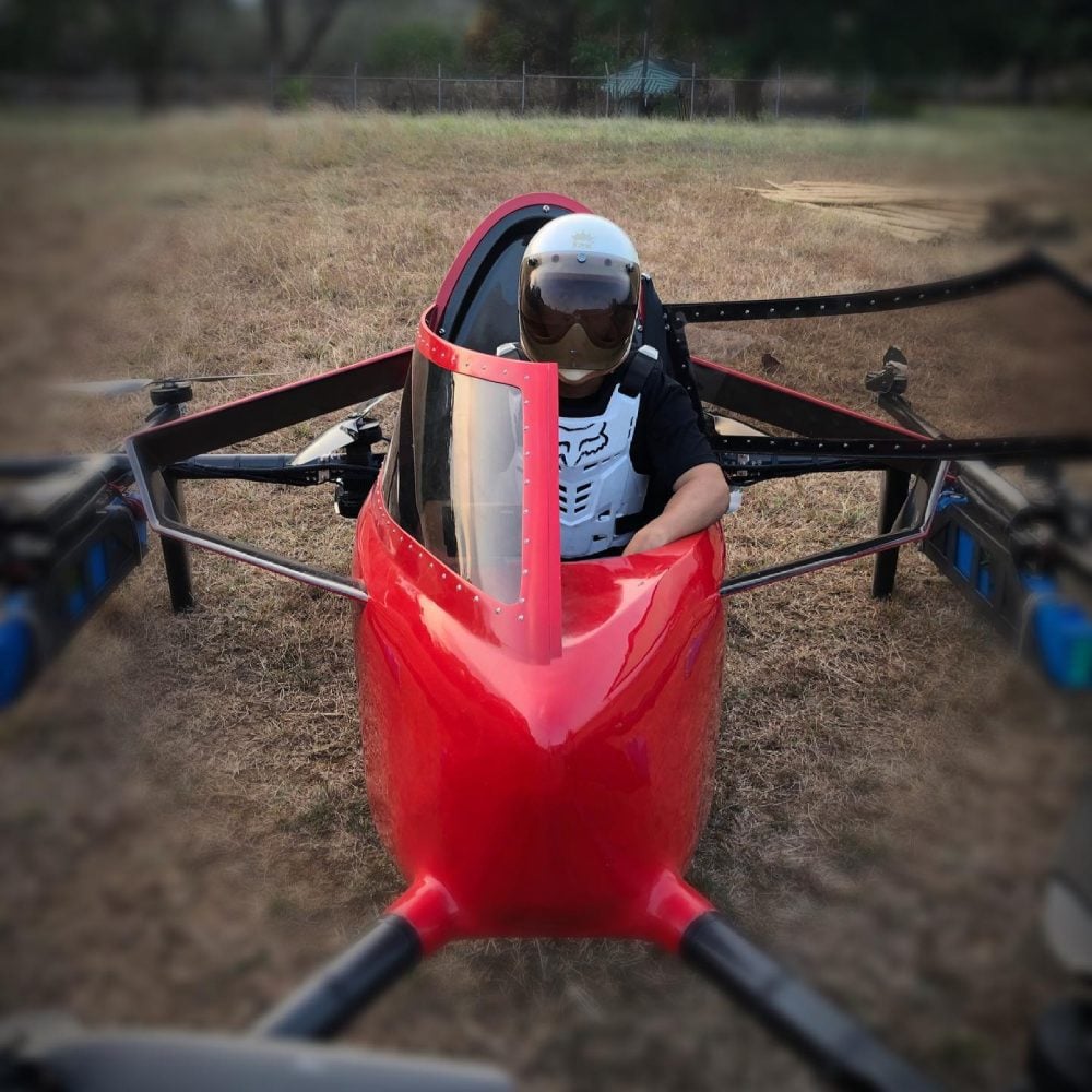 Pilot sits in a red single-seat multirotor flying car on dry grass.