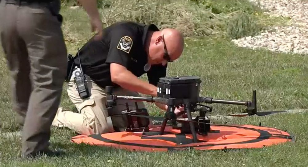 Officer kneels to prepare a drone on an orange landing pad in grass.