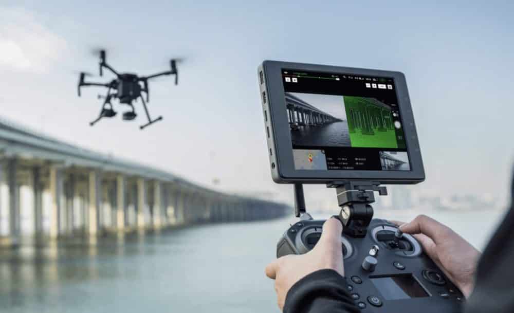 Pilot holding a controller and monitor while a drone inspects a long bridge.