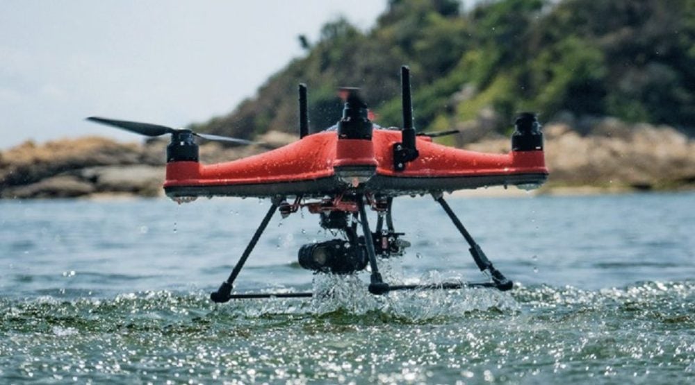Red SwellPro SplashDrone 4 lifting out of the sea near a wooded shoreline.