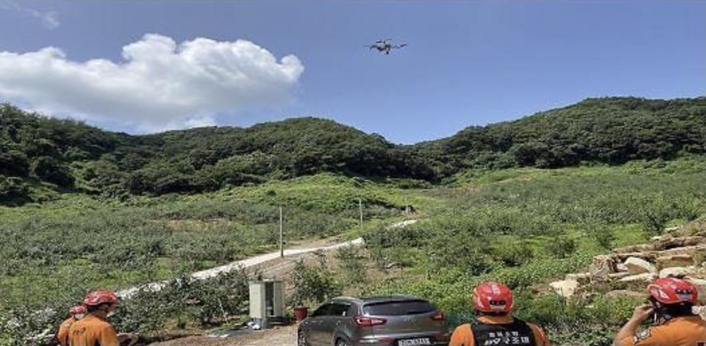 Drone flies above a hillside track as rescuers gather near a parked car.