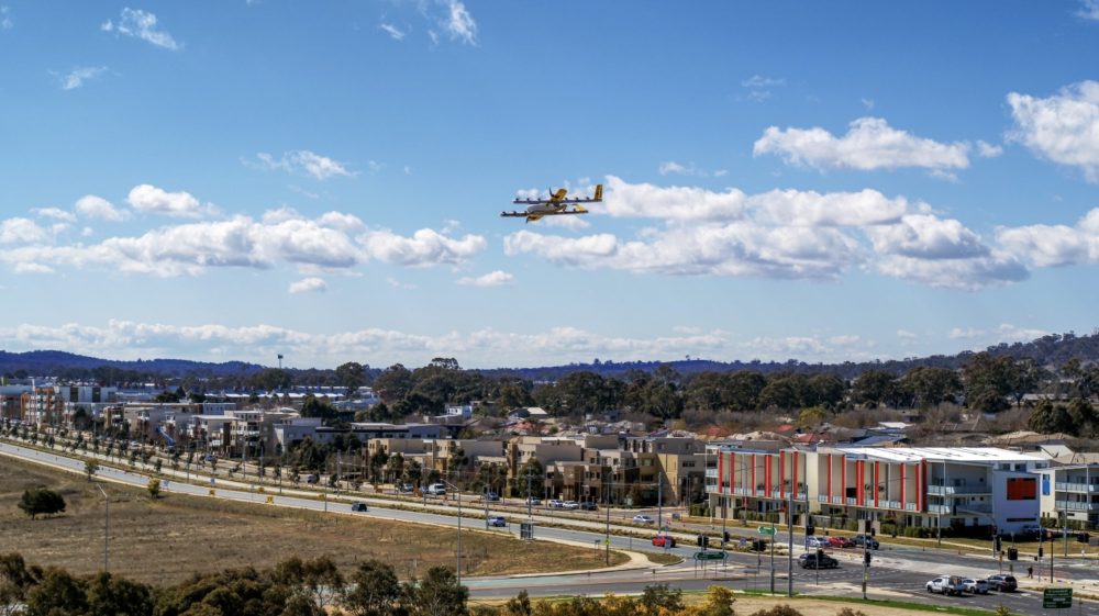 Wing delivery drone hovering above a suburban Australian town with wide arterial roads.