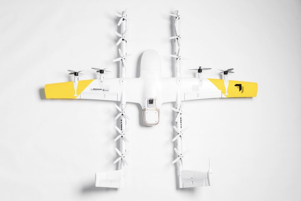 Overhead view of a Wing delivery aircraft with yellow wingtips and a hanging package.