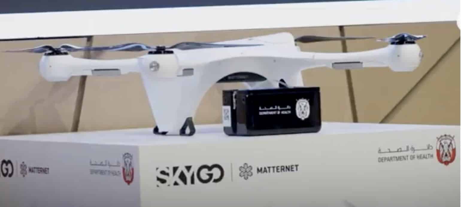 A white Matternet delivery drone with payload box in front of SkyGo and Matternet banners.