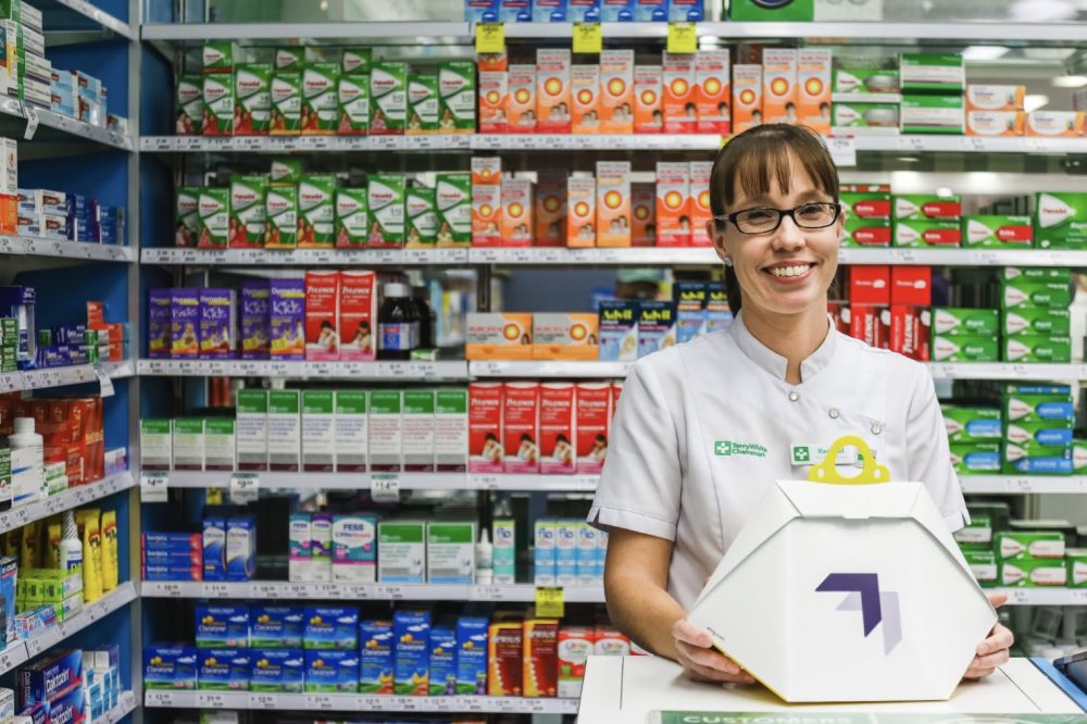 Pharmacy worker holds a hexagonal drone delivery package before medicine shelves.