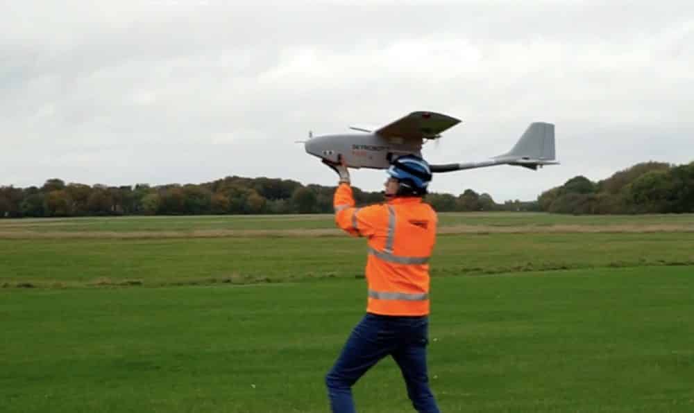 Operator in a high-visibility vest hand-launches a fixed-wing drone over a field.