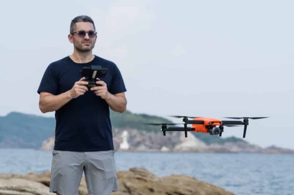 A pilot holds a controller as an orange Autel drone hovers above a rocky coastline.