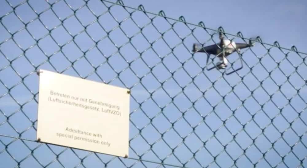 Drone hovering behind a chain-link fence beside a German restricted-access sign.