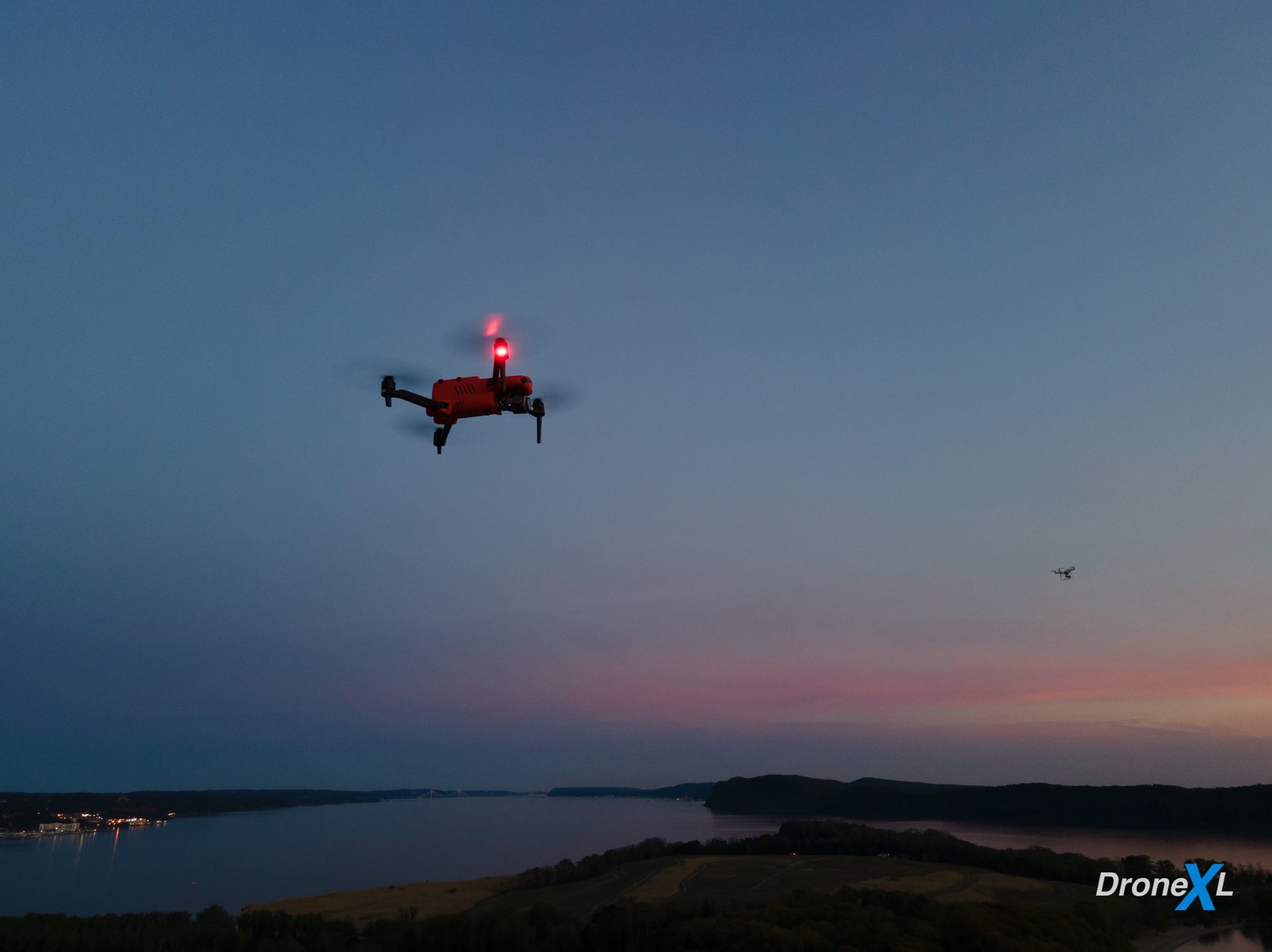 Drone hovering at dusk above a river valley with its red light lit.