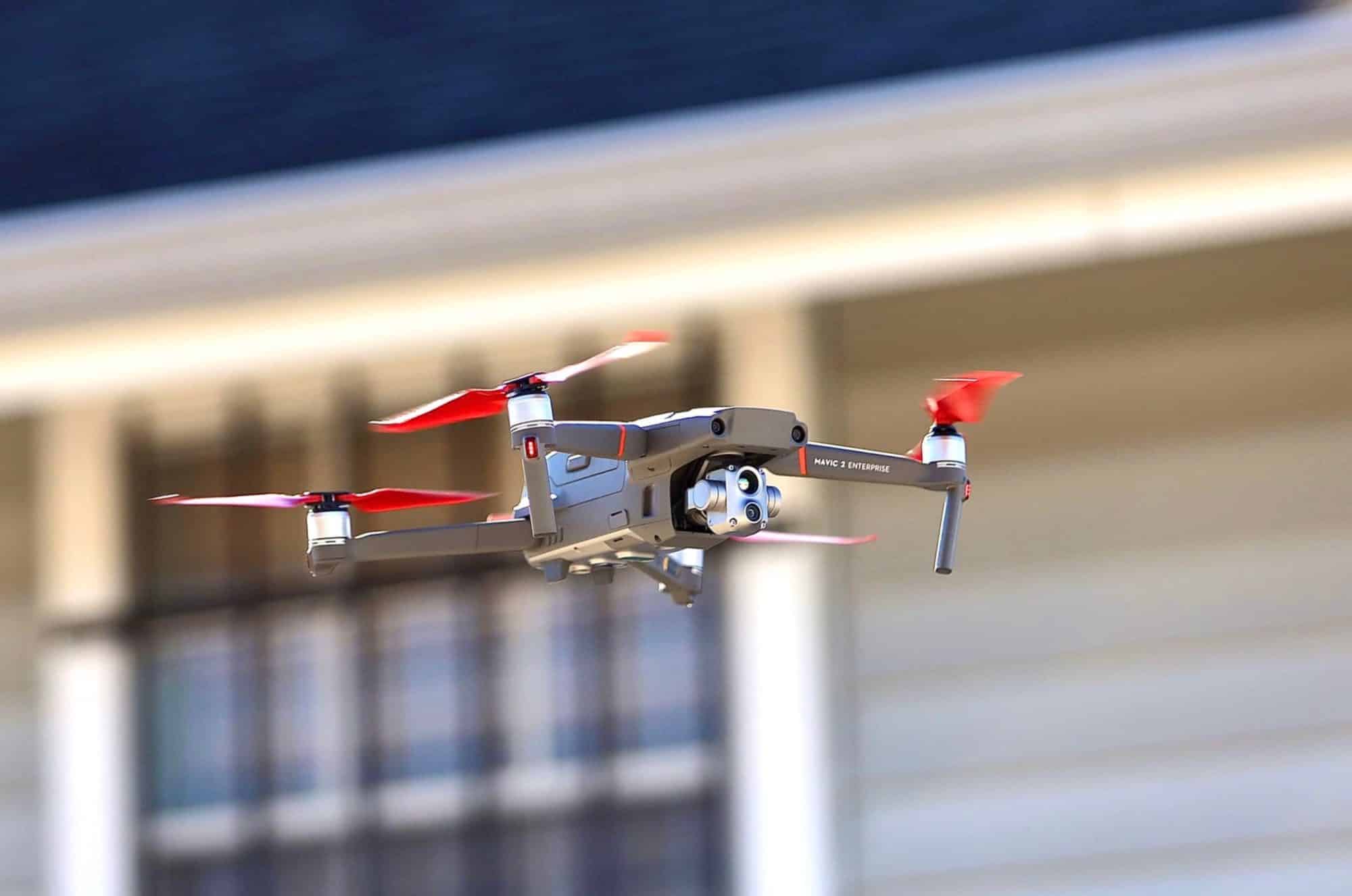 DJI Mavic 2 Enterprise Advanced with red propellers flying past a building facade.