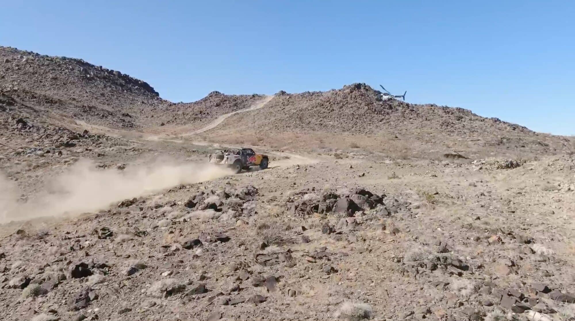 Drone footage of an off-road race car trailing dust with a helicopter beyond the ridge.