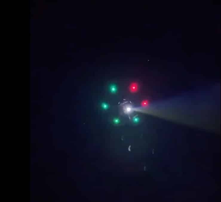 Drone hovers in darkness with green and red navigation lights and a bright spotlight.