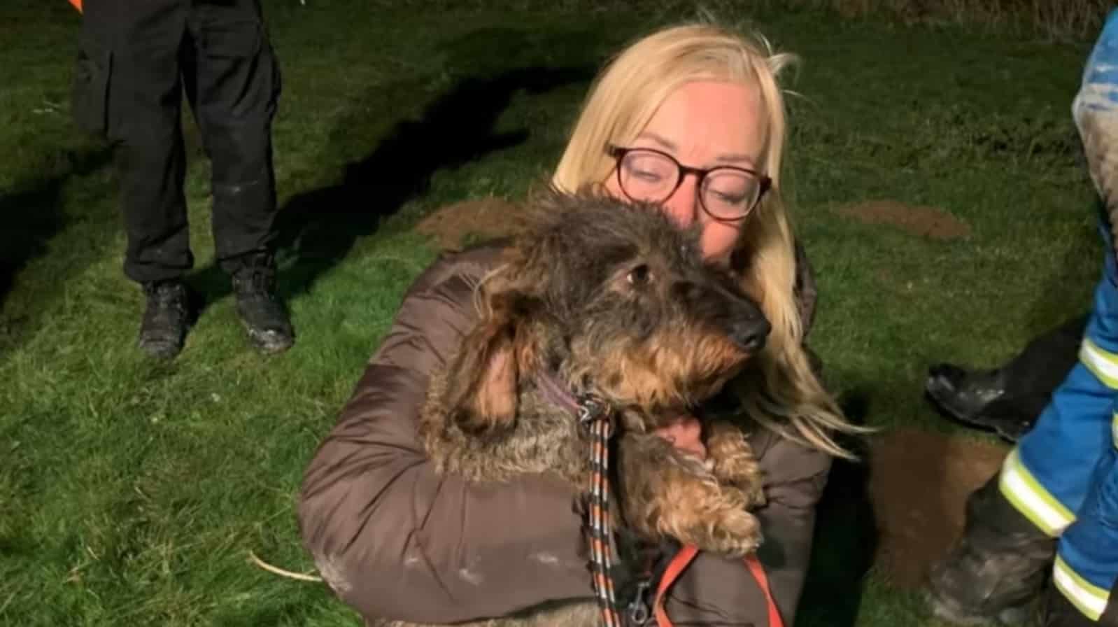 Owner cradling the rescued wirehaired dachshund as firefighters stand nearby.