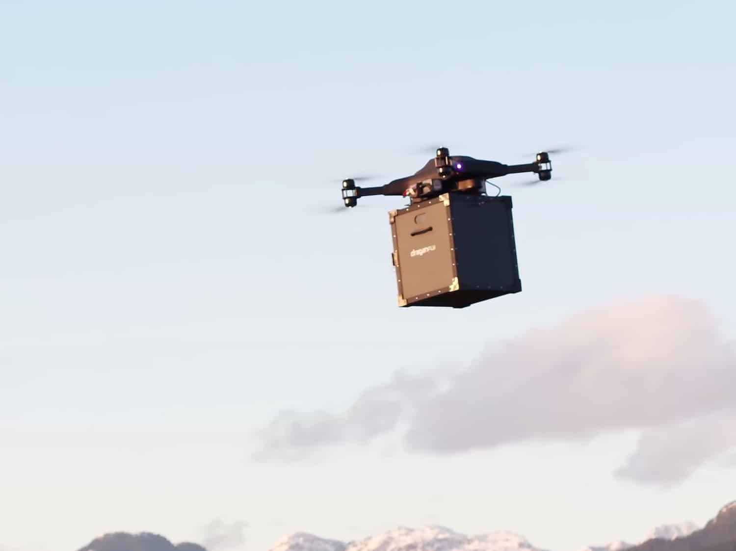 A delivery drone carrying a cardboard box aloft against a pale mountain sky.