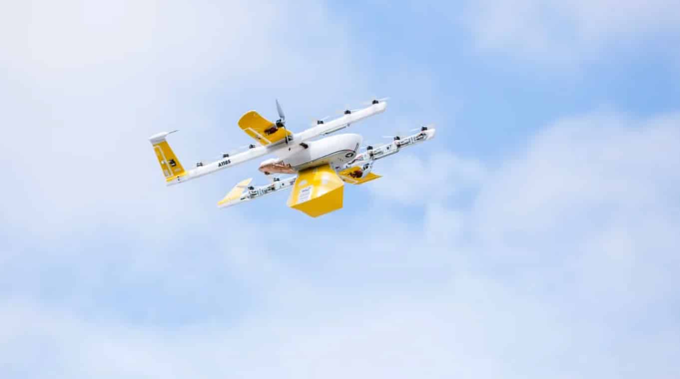 Wing delivery drone climbing with a yellow package clipped beneath it.