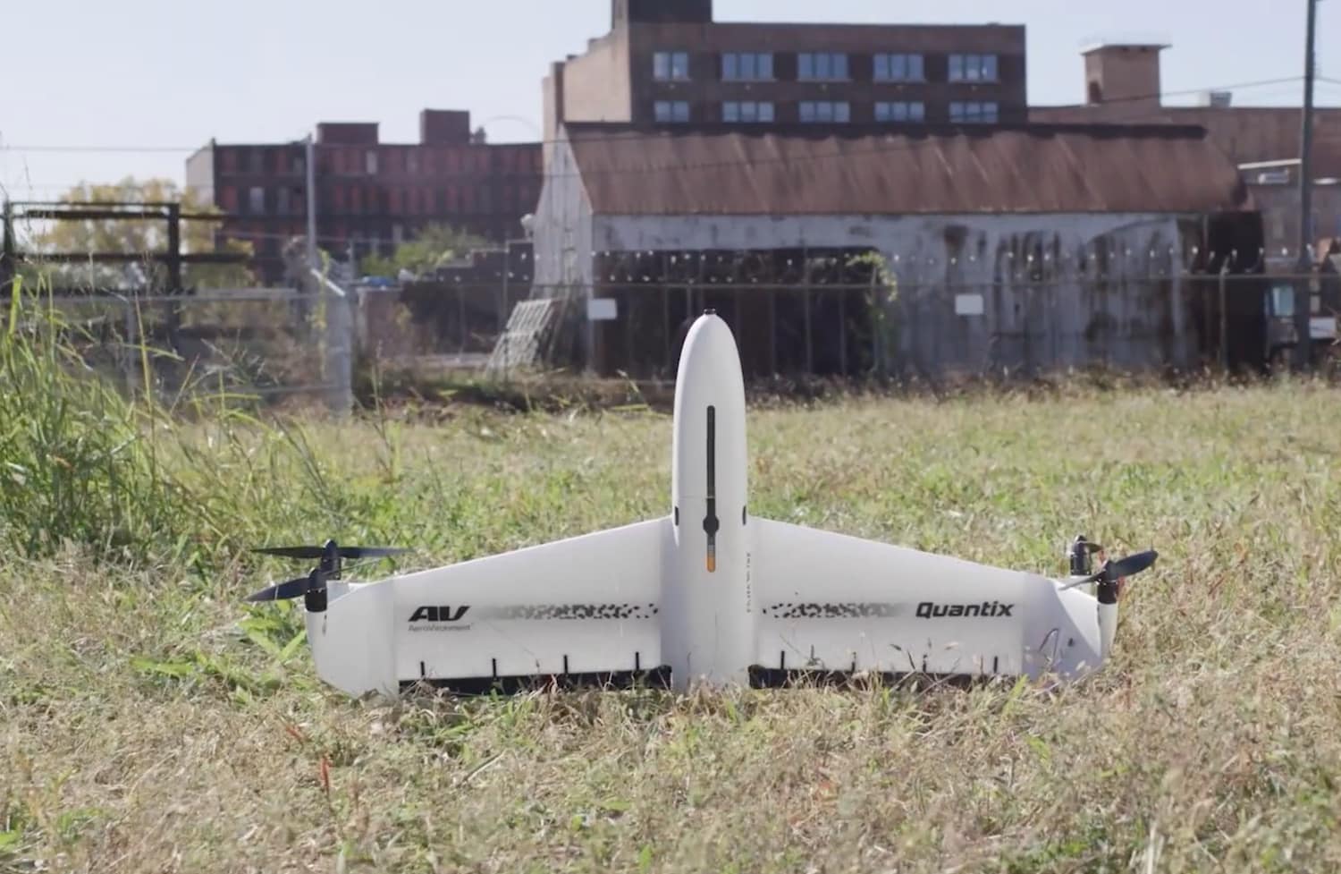 AeroVironment Quantix Recon fixed-wing drone resting on grass beside derelict industrial buildings.