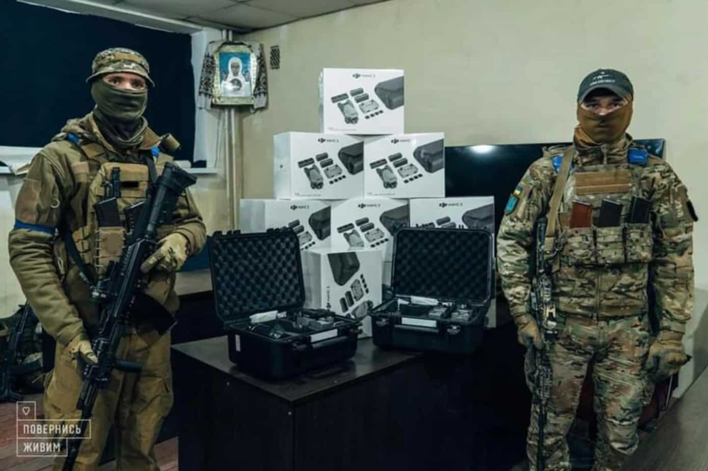 Two masked soldiers stand beside stacked DJI drone boxes and hard cases indoors.