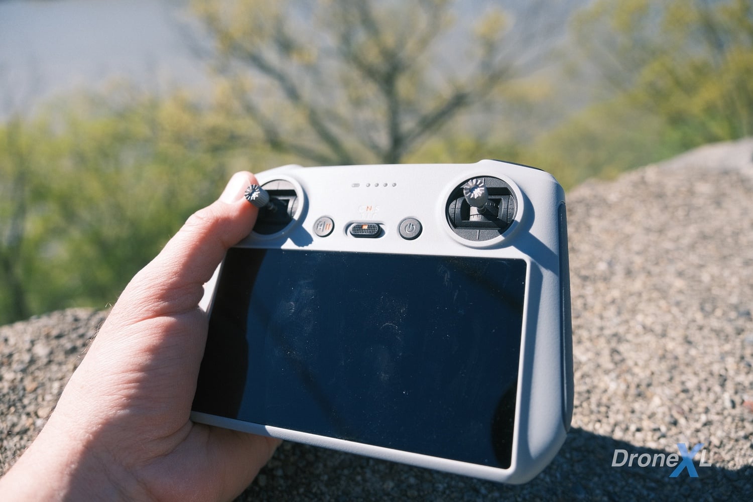 A hand holds a DJI RC screen controller outdoors above a gravel ledge.