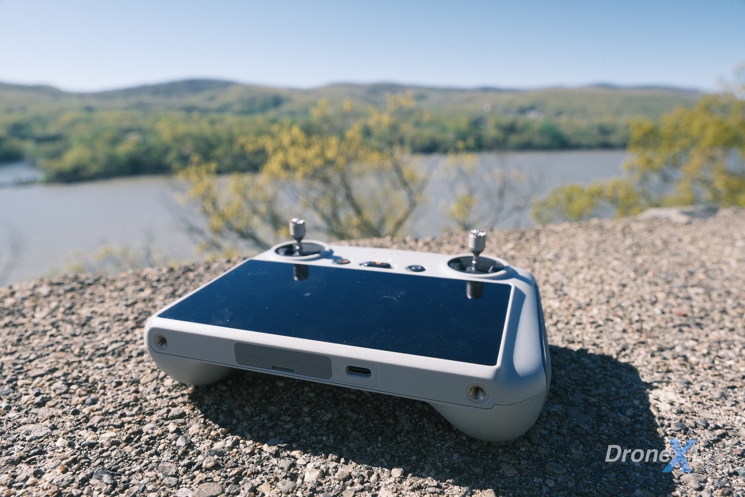 DJI RC controller resting on gravel with a river and wooded hills behind.
