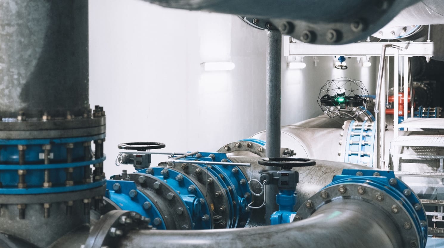An Elios 3 caged inspection drone flying among blue industrial pipes and valves.