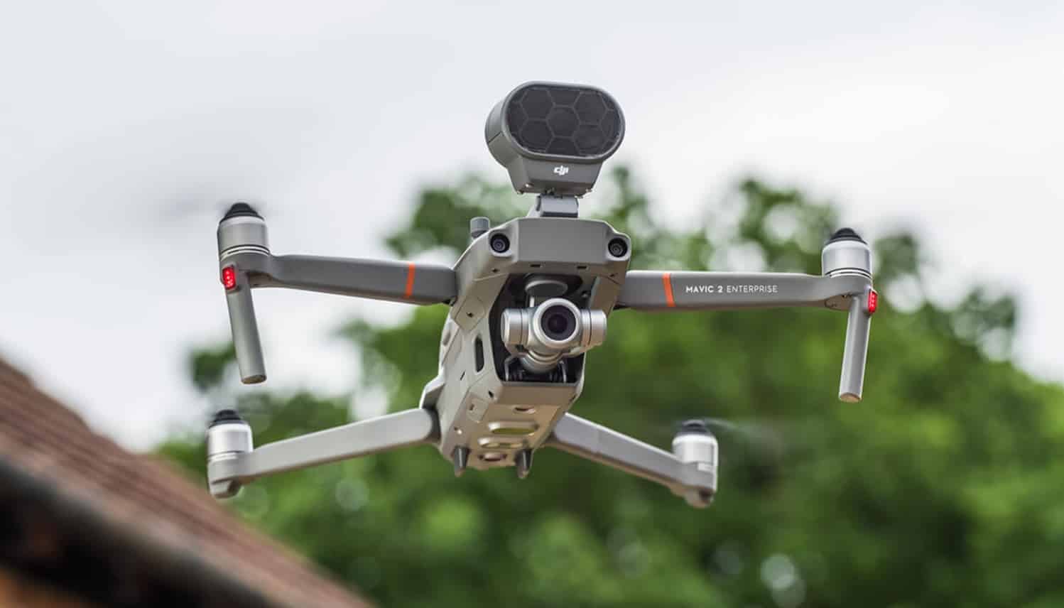 DJI Mavic 2 Enterprise carrying a loudspeaker module flying past a rooftop.