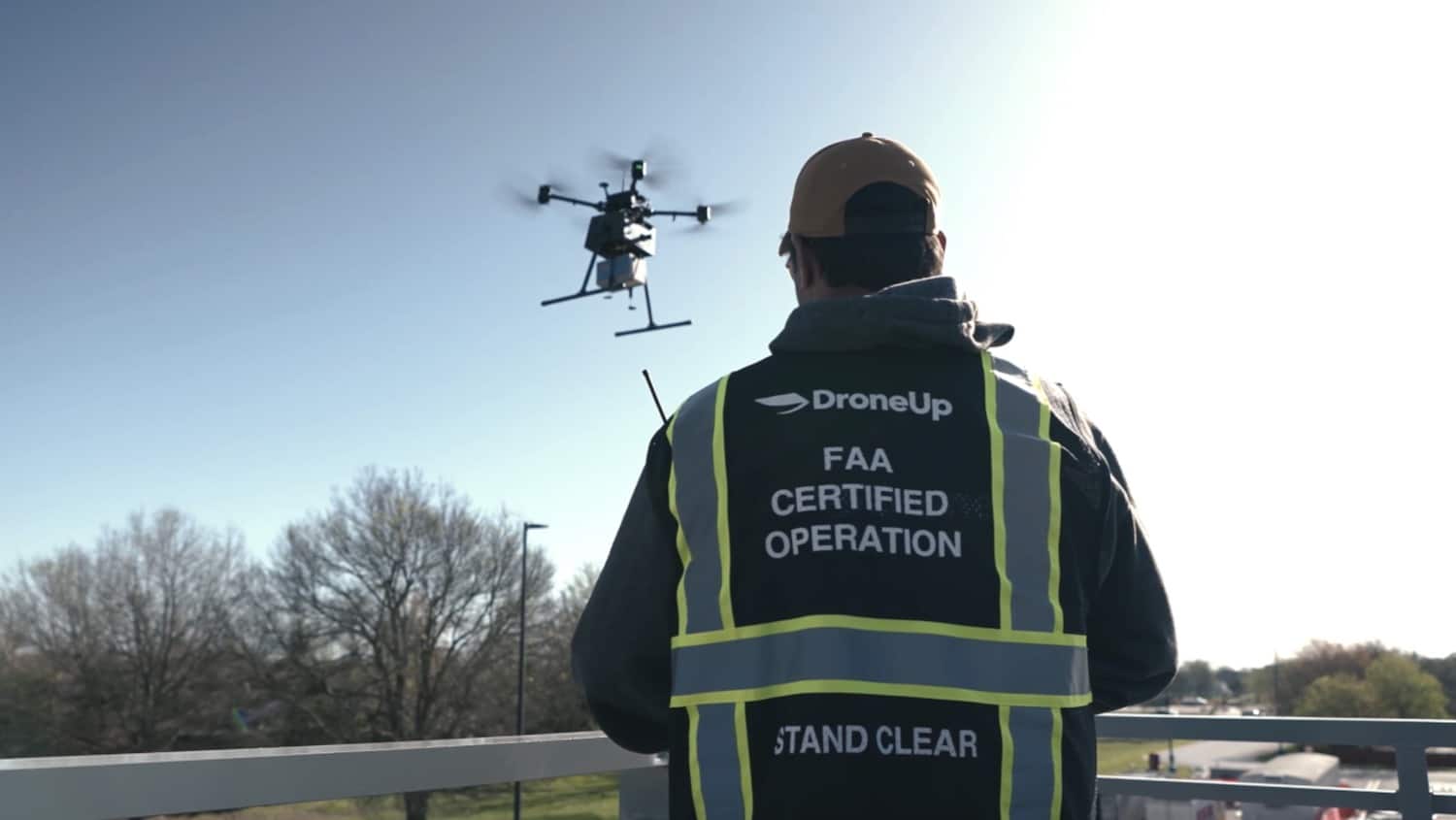 DroneUp operator in a certified operation vest watching a delivery drone overhead.