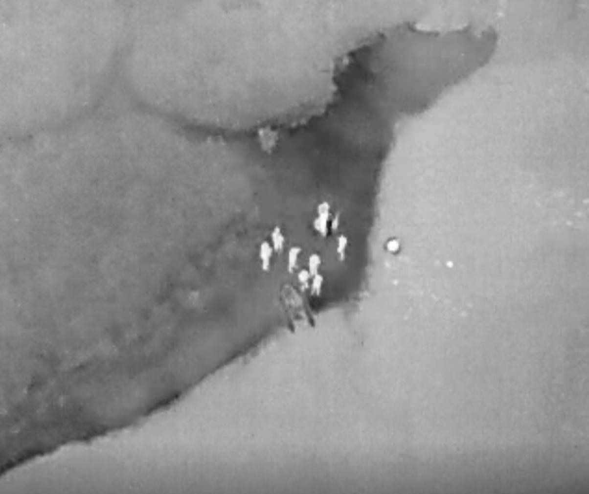 Thermal aerial view of firefighters gathered at a riverbank with a small inflatable boat.