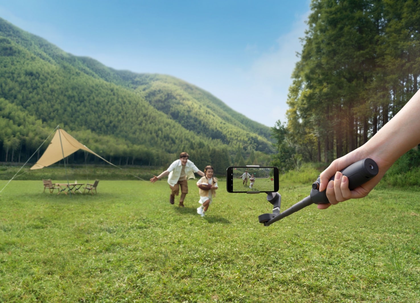 A hand holds the Osmo Mobile 6 while filming two people running across a campsite meadow.
