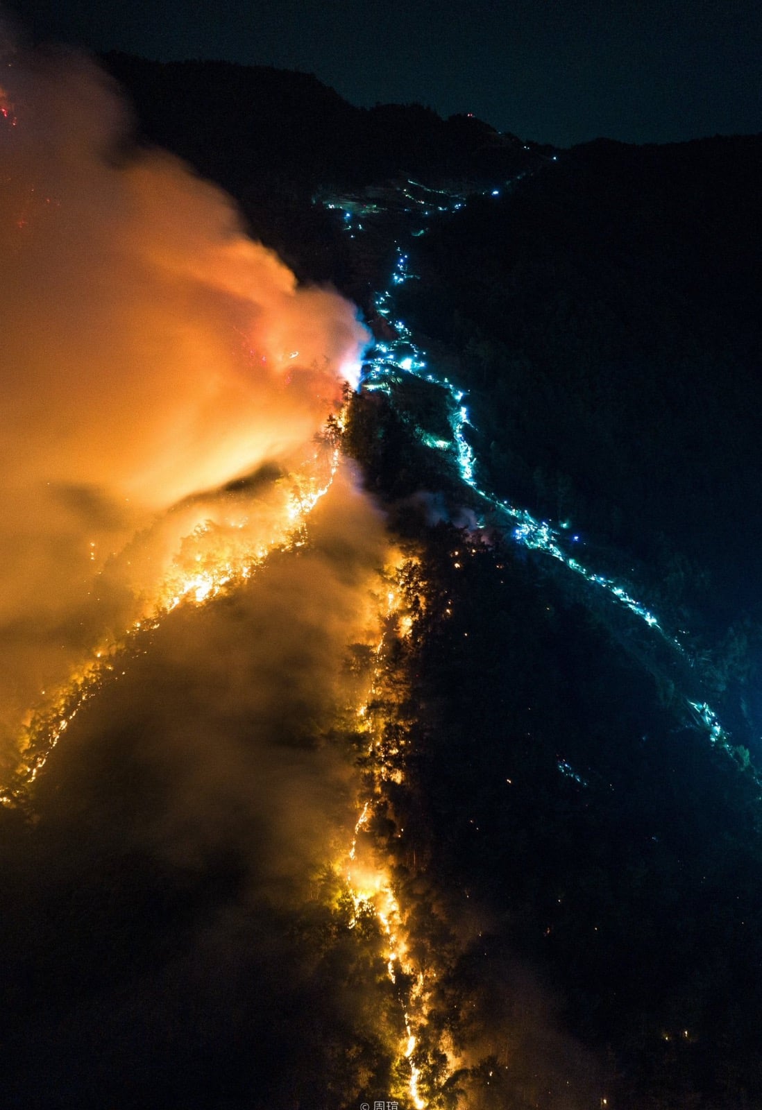 Night aerial view of a burning ridgeline with a blue chain of lights alongside.