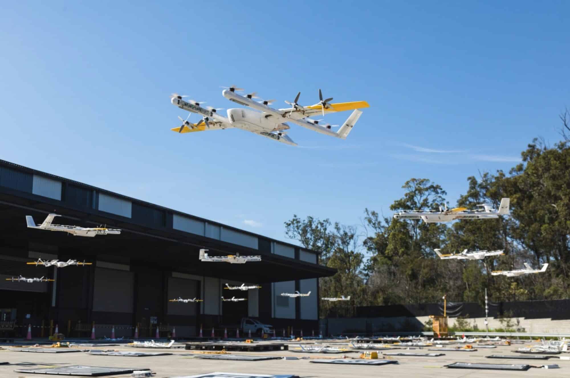 Wing delivery drones hover above a store rooftop launch pad.