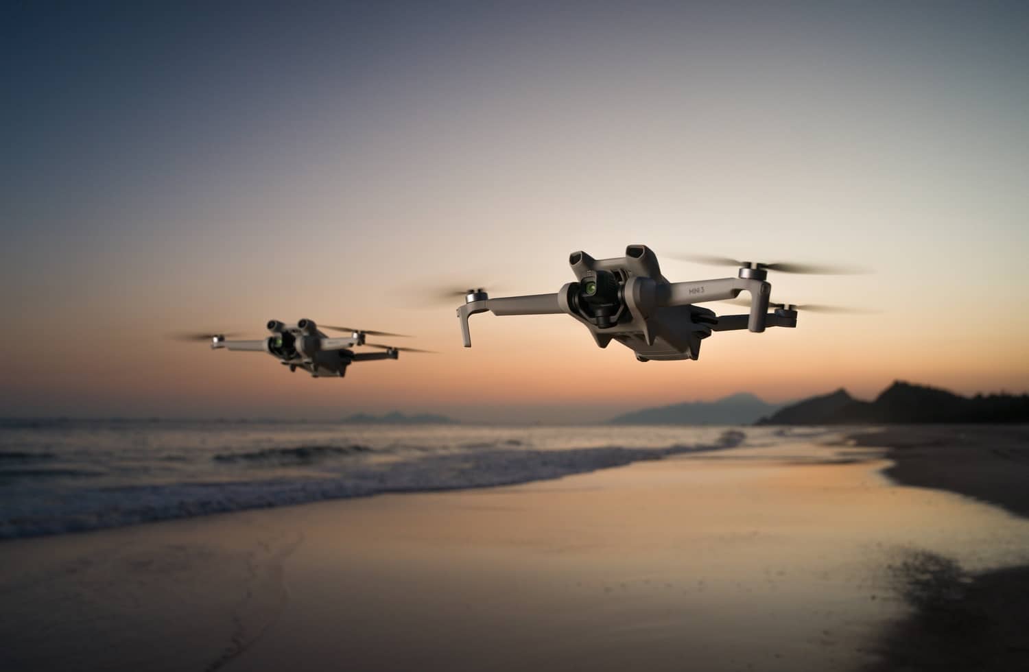 Two DJI Mini 3 drones fly over a beach at sunset.
