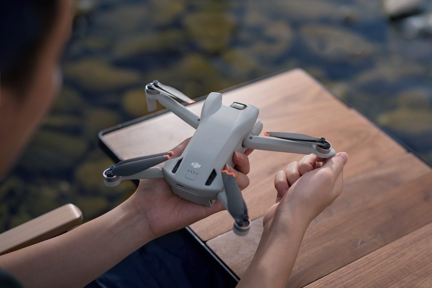 Hands hold an unfolded DJI Mini 3 above a wooden table beside a stream.