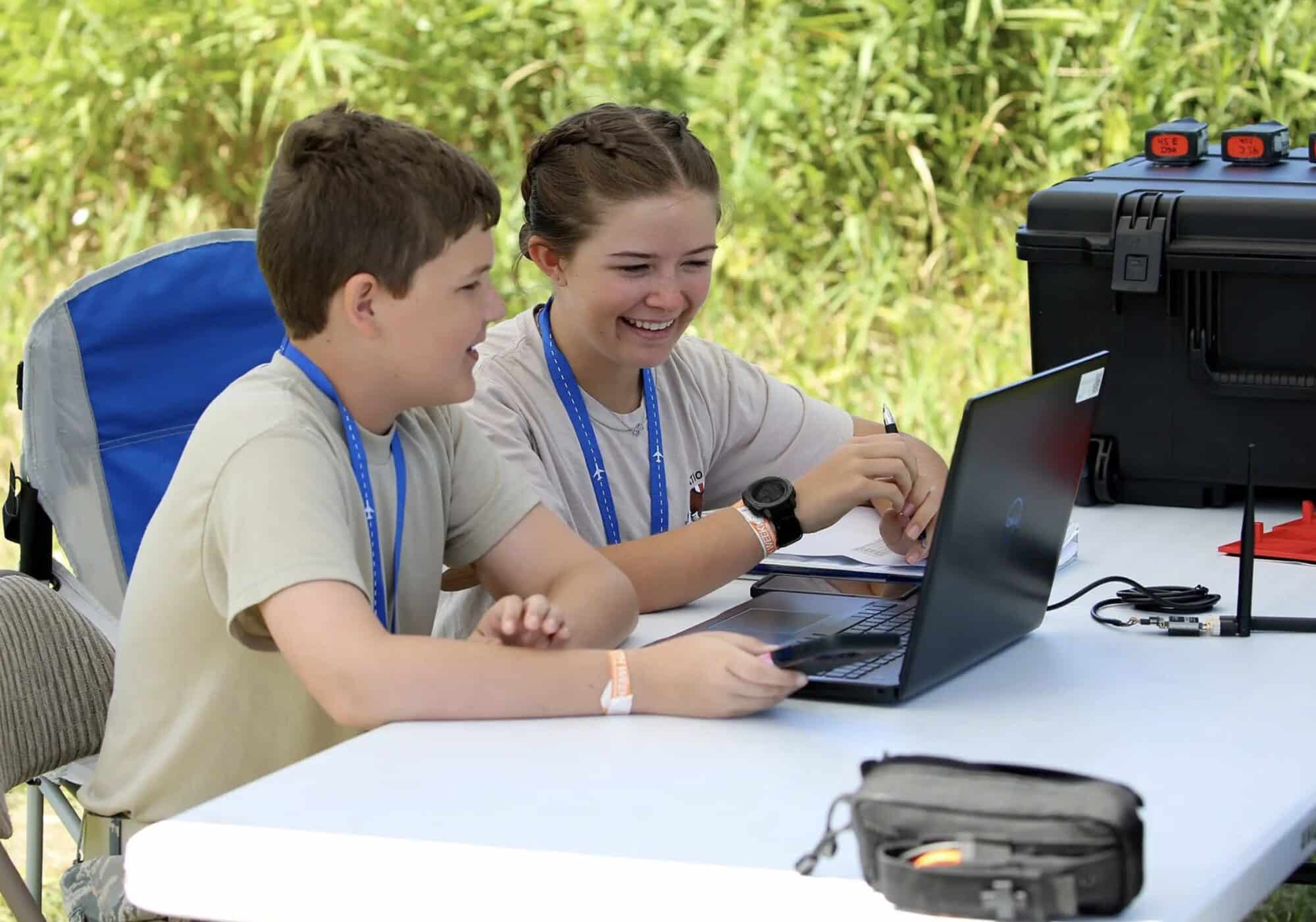 Two teenage competitors sharing a laptop at an outdoor drone competition table.