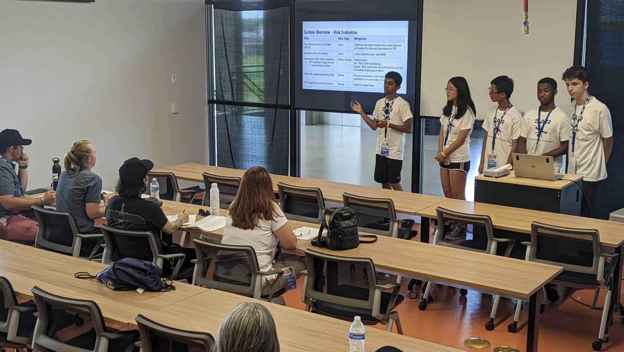 Student team presenting to a seated audience in a classroom with a slide.
