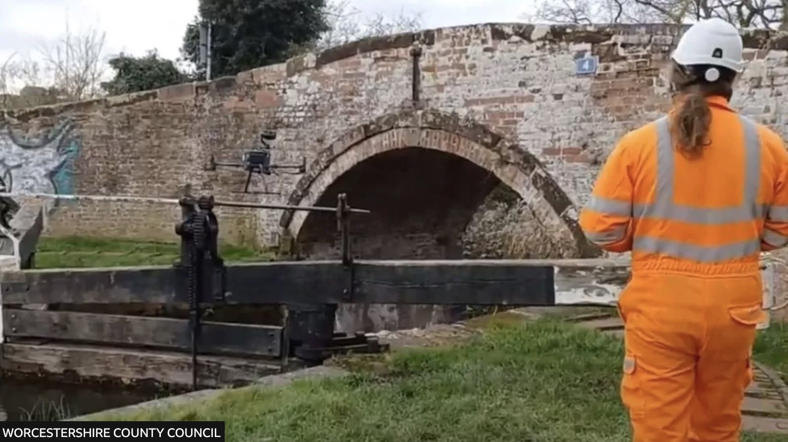 Worker in orange high-visibility gear inspecting an old brick arch bridge by drone.