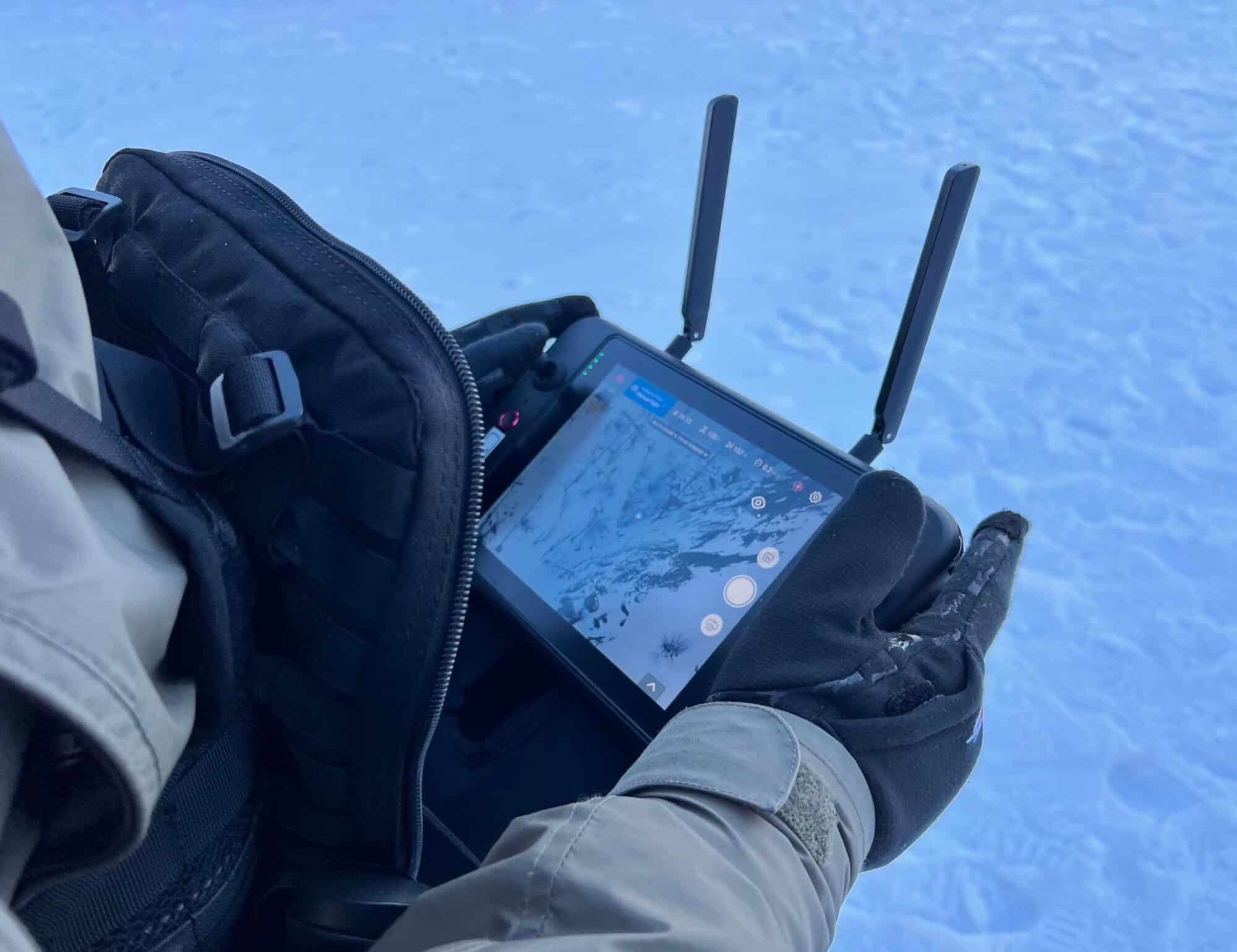 Gloved hands hold a drone controller showing a map screen above snowy ground.