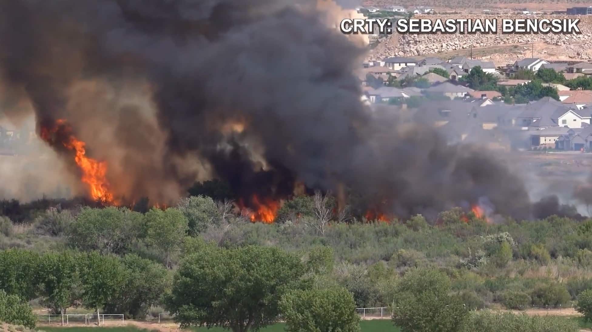 Flames and black smoke rise from riverside brush below Washington City homes.