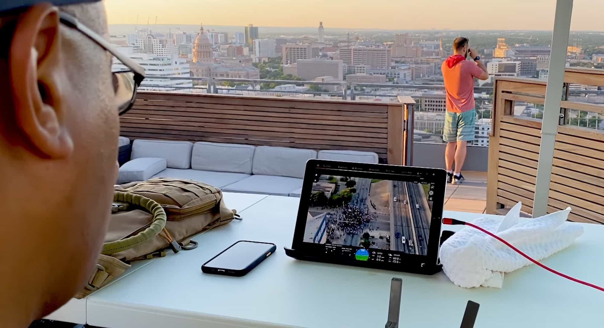 Officer on a rooftop watching a tablet showing a drone live feed downtown.