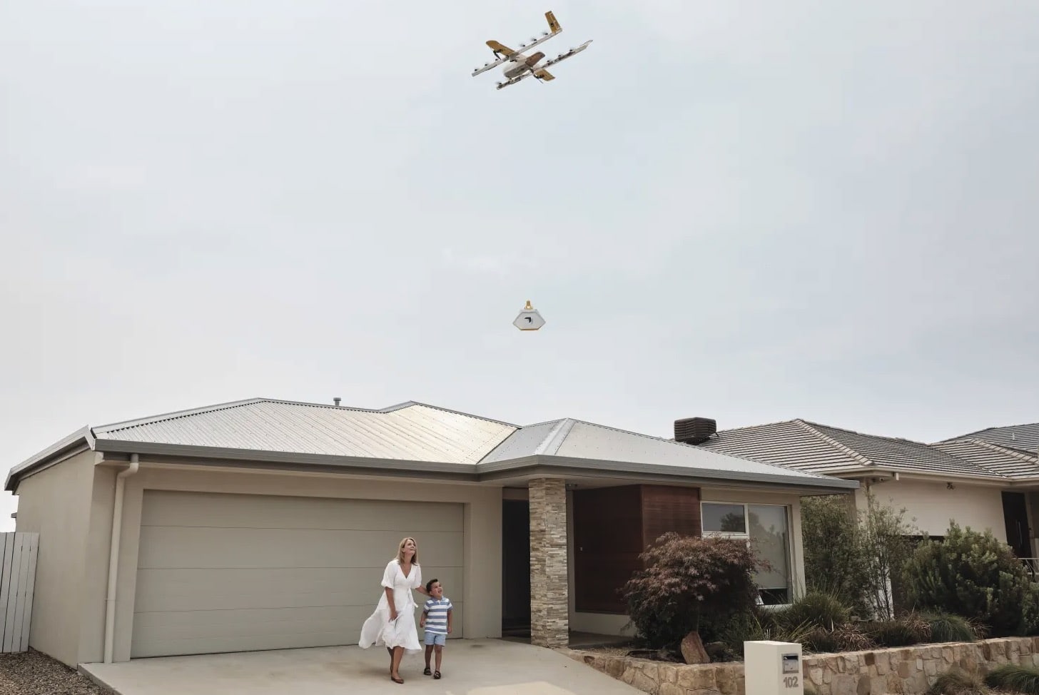 Wing drone lowering a package on a tether to a family outside their home.