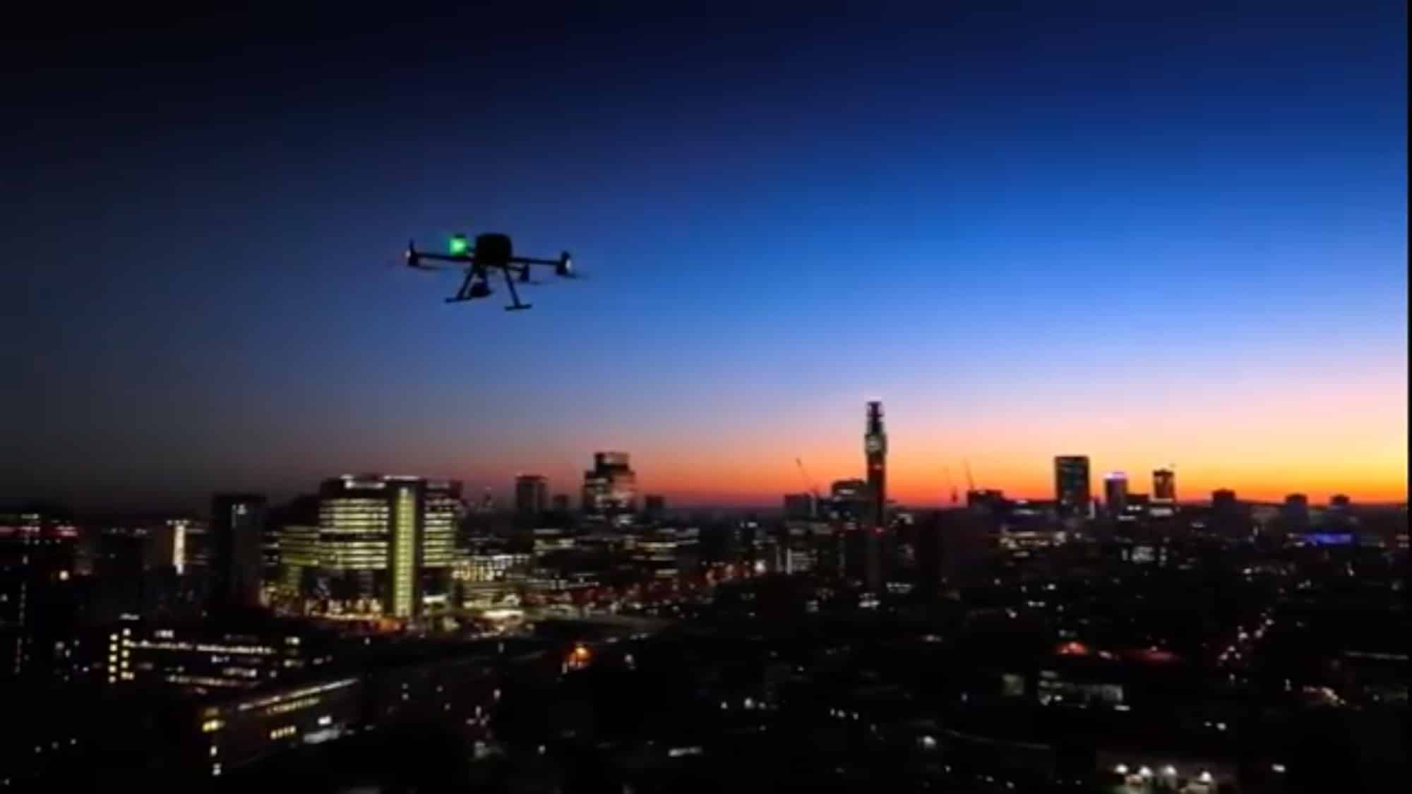 Police drone hovers above a city skyline at dusk as lights come on.
