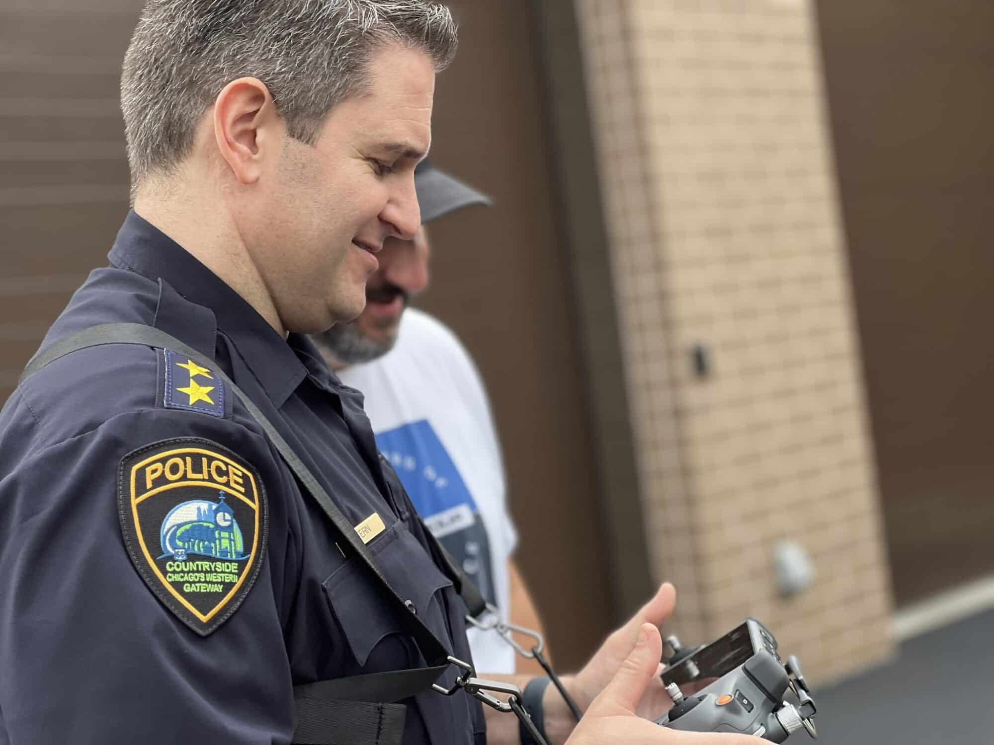 Countryside Police officer watches a colleague operate a drone controller outside a building.