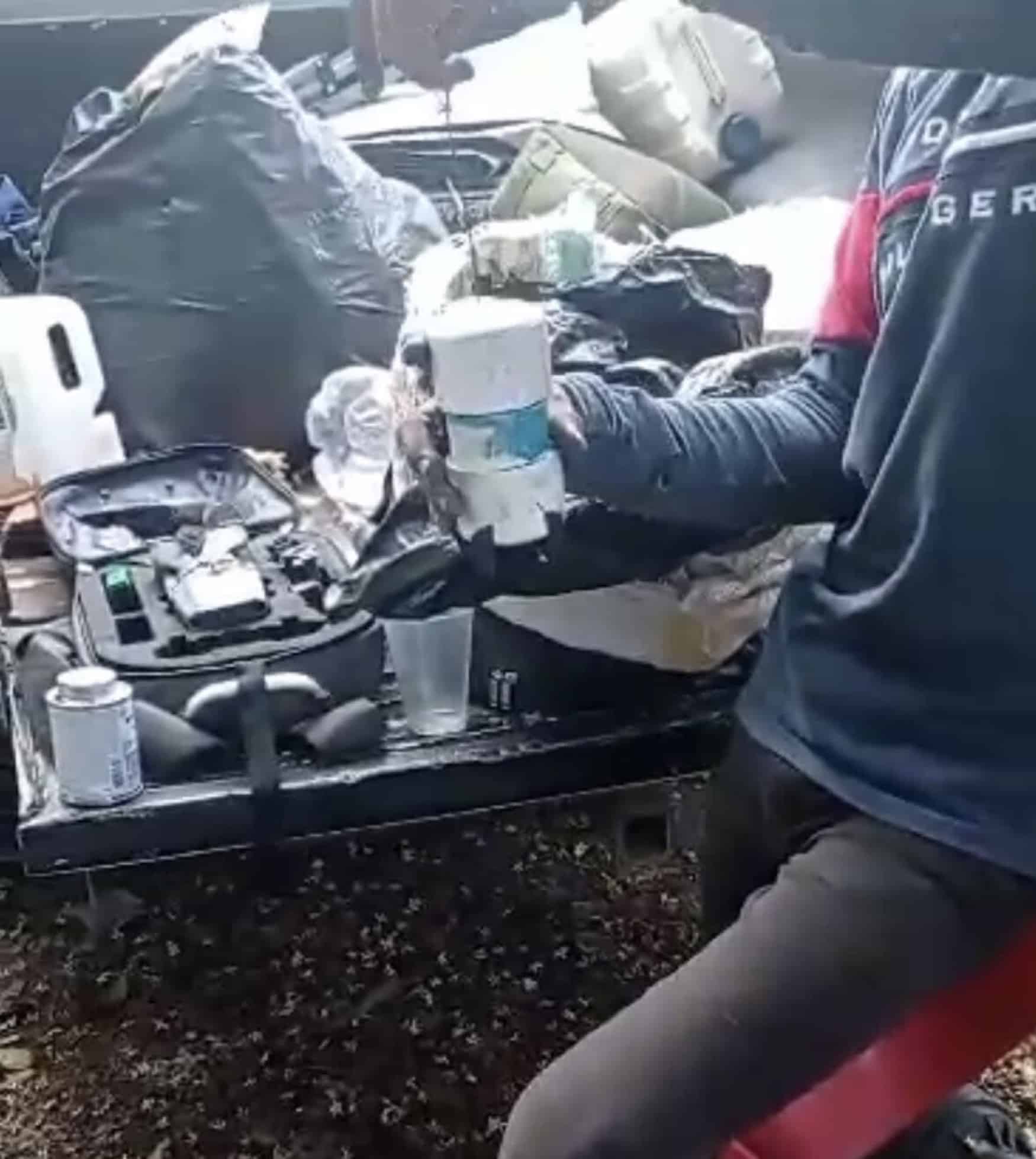 Video still of a person rigging explosives into plastic bottles beside a drone case.