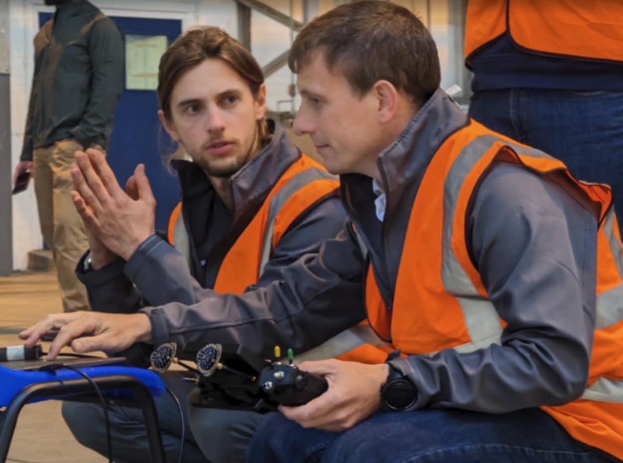 Two engineers in high-visibility vests operate drone controllers during the test flight.