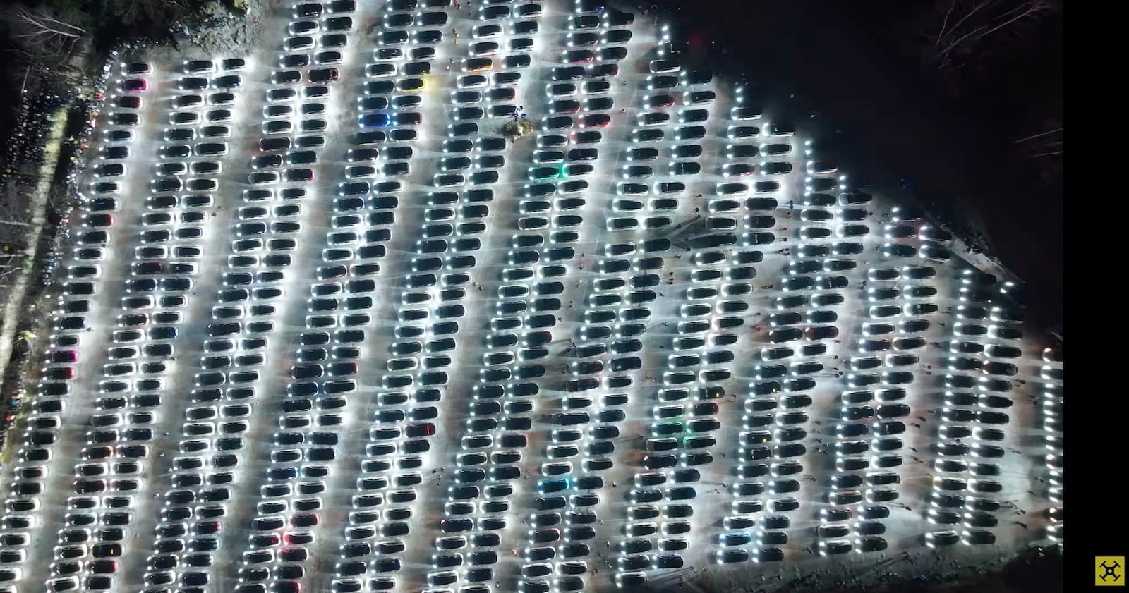 Aerial night view of hundreds of parked Teslas flashing their lights in formation.